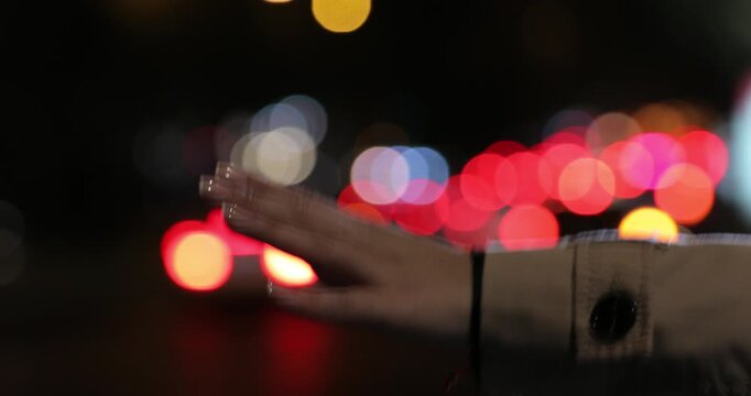 Close up shot of a woman's hand in a trench coat waving to hail a cab against a beautifully blurred background of colorful city traffic lights at night.


