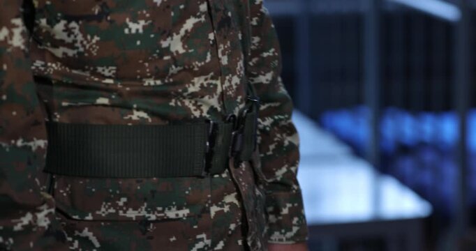 Close-up shot of an anonymous soldier in a digital camouflage uniform carefully fastening a wide, dark green tactical belt around their waist against a softly blurred indoor background.

