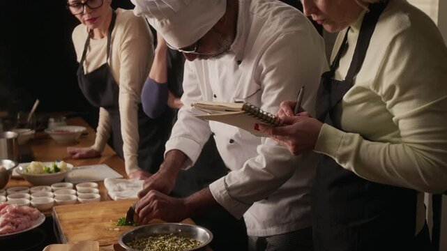 Close up over the shoulder shot of woman taking notes in notebook while attending group cooking master class with ither women in restaurant