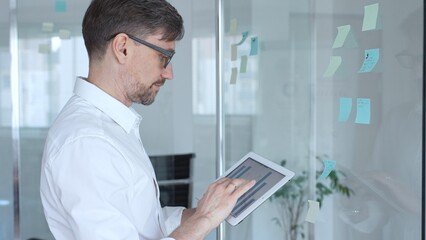 Fototapeta premium Business person analyzing data, brainstorming new strategies using digital tablet and adhesive notes on a glass board, planning future project development during an office meeting