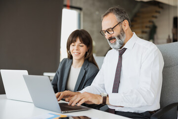 Fototapeta premium A senior professional man and a young female colleague working side-by-side on their laptops in a modern office