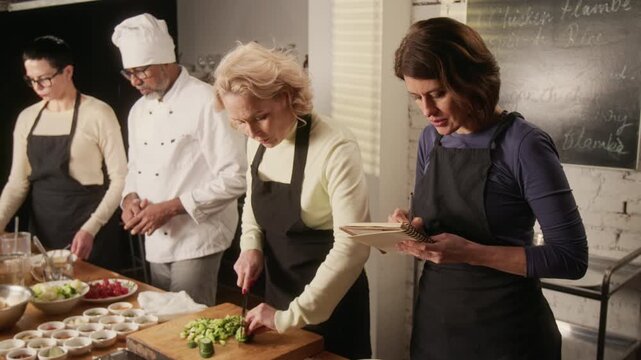 Woman taking notes in notebook, another chopping vegetables, man in chefs hat watching, and third woman washing dishes during cooking master class