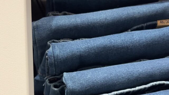 Close up video. 60 fps. Camera moves along shelf with new blue jeans pants, denim clothes, jackets, shirts, dress, textile thing, overalls in clothing store. Concept of 11 november sale, shop, promo