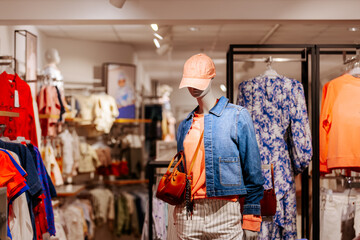 Obraz premium Female mannequin dressed in denim jacket, orange sweater, and striped pants stands in a clothing store with various garments displayed on racks in the background