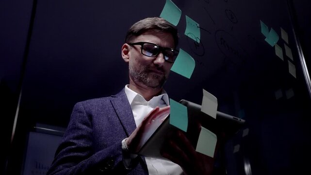 Mature male professional, wearing eyeglasses and suit, working late in a dark office, holding a digital tablet and brainstorming new business strategies with post it notes on a glass board