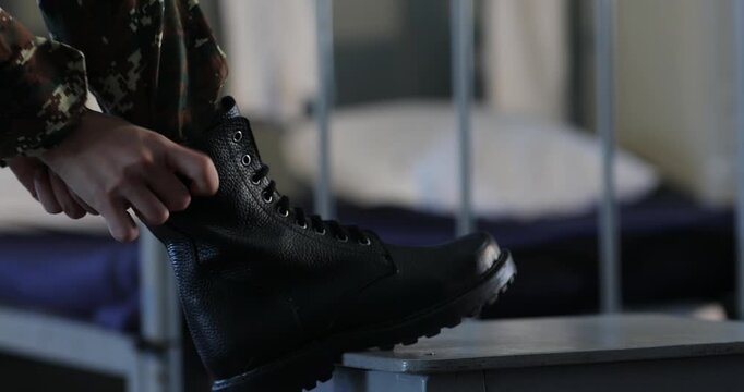 An unrecognizable soldier wearing camouflage pants is carefully tying the shoelaces of heavy black combat boots while resting one foot on a small stool in a barracks room.

