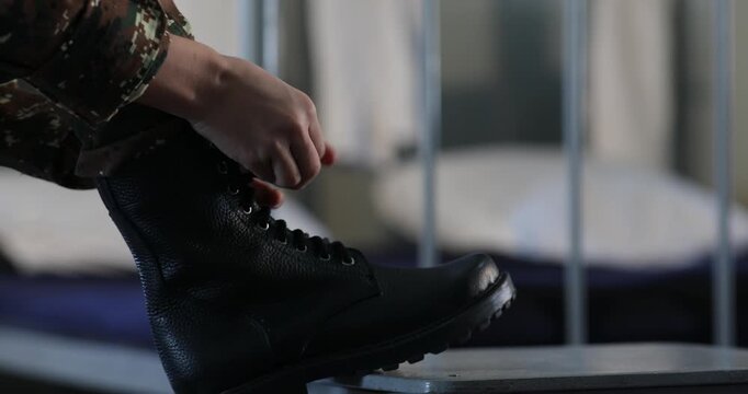 An unrecognizable soldier wearing camouflage pants is carefully tying the shoelaces of heavy black combat boots while resting one foot on a small stool in a barracks room.

