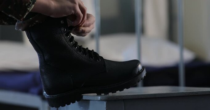 An unrecognizable soldier wearing camouflage pants is carefully tying the shoelaces of heavy black combat boots while resting one foot on a small stool in a barracks room.


