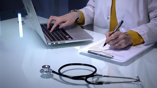 Stethoscope laying on a white reflective desk while medical professional working in clinic office. Typing on laptop and writing notes. Medicine concept