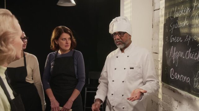 Middle aged biracial male chef discussing menu on chalkboard with group of women, attending restaurant cooking master class