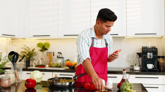 Indian man frying vegetables in pan while checking product list on smartphone for healthy meal prep in home kitchen. Arabian guy focused on diet routine and fresh food choice for balanced nutrition.
