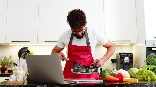 Arabian man tosses vegetables in frying pan while checking laptop recipe tips in kitchen for healthy meal prep. Young guy stays focused on home cooking for diet fitness nutrition in apartment indoors.