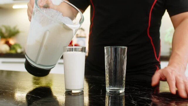 Man pours protein smoothie from blender into glass on kitchen counter for healthy nutrition. Shake with banana milk cottage cheese and whey protein supports muscle recovery weight control fitness gym.