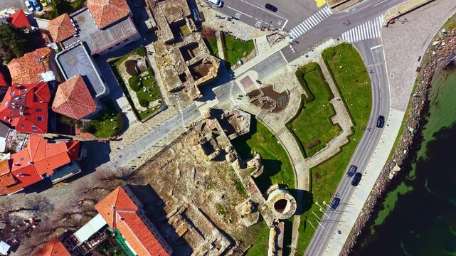 Nesebar Old Town shows ancient ruins and narrow streets with red-roofed buildings. Visitors walk near the coast enjoying a sunny day and the view of the sea.