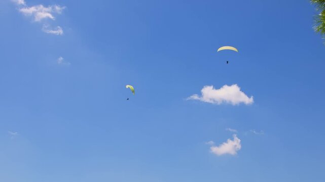 Two paragliders perform a tandem flight high above the Turkish coast against a bright blue summer sky with white fluffy clouds. This cinematic footage captures the essence of extreme sports, freedom, 