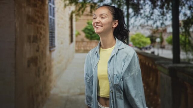 Young hispanic woman clapping hands, smiling and looking aside on a sunlit stone street beside a carved balustrade and building wall; joy celebration.