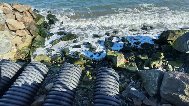 Waves hit the shore as rocks line the beach. Drainage pipes are placed in front of the water. Bright sunlight illuminates the scene and shows the natural elements at play.