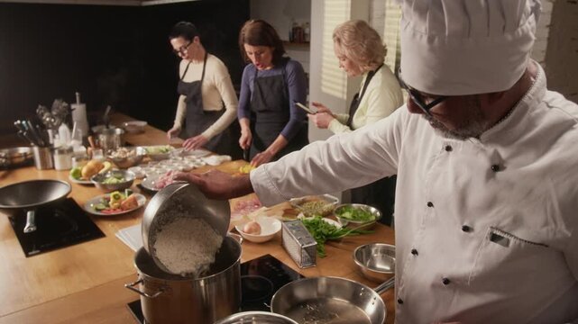 High angle tilt down zoom in shot of biracial middle aged chef cooking rice while group of women cutting vegetables during cooking master class in restaurant