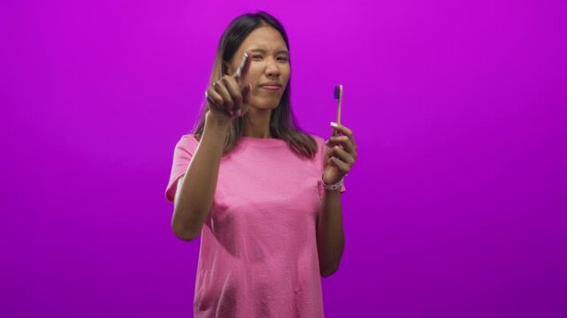 Woman holds a toothbrush in her right hand while pointing index finger to camera and making an l gesture at forehead in studio with vibrant purple backdrop; oral hygiene confidence.