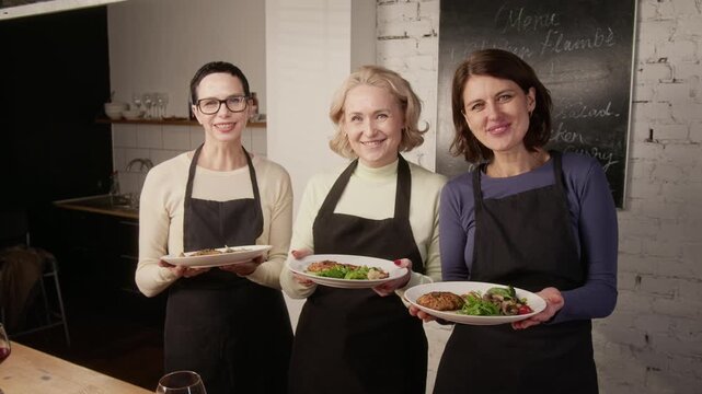 Portrait shot of three smiling women in dark aprons standing with plates of food in restaurant kitchen with chalkboard
