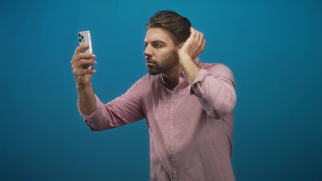 Hispanic man with beard in a pink button shirt holds a smartphone at arm's length and runs his hand through hair while checking his face in a blue studio; grooming confidence.