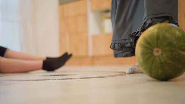 White girl stands near green squash on floor, patterned socks and long skirt step across warm wooden kitchen, playful motion toward produce, sibling scene suggesting kitchen cook preparation, soft