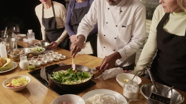 High angle tilt down shot of chef adding spinach to rice in pan during group cooking master class in restaurant
