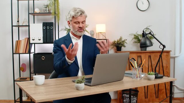 Middle-aged businessman at home office teaches live webinar via laptop wearing earphones explaining concepts. Freelancer guy at table replies to user questions giving practical examples for training.