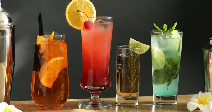 Different tasty refreshing cocktails in glasses, shakers and jigger on wooden table against gray background. Camera moving right