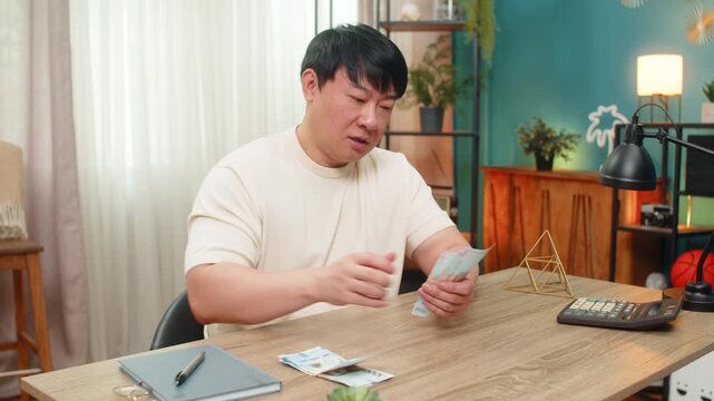 Asian man at table counts UAH Ukrainian hryvnia cash bills, sets part of money aside, showing careful financial discipline. Chinese guy keeps satisfied expression records totals planning budget calmly