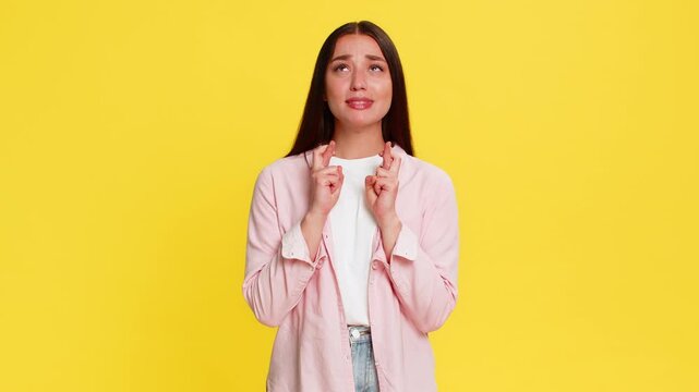 Young woman crossing fingers and closing eyes with hopeful expression, silently wishing for luck and good outcome. Girl on yellow background shows emotional anticipation and sincere desire for success