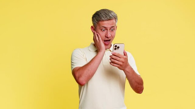 Middle-aged man scrolling smartphone then suddenly freezes in shock, with wide eyes and mouth open, stunned by breaking news. Young guy reacting to unexpected message isolated on yellow background.