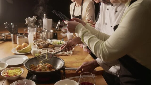High angle tilt down shot of professional chef frying meat and vegetables in pan while woman filming it with smartphone during cooking master class