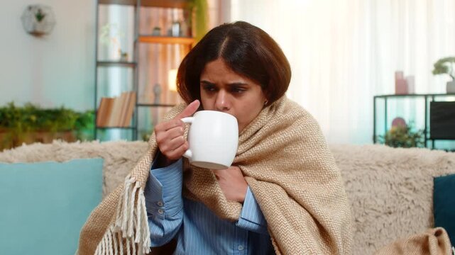 Indian woman coughs and drinks tea under blanket on home sofa, showing flu symptoms and low energy. Arabian girl appears unwell, tired, with facial expression of weakness and seasonal discomfort.