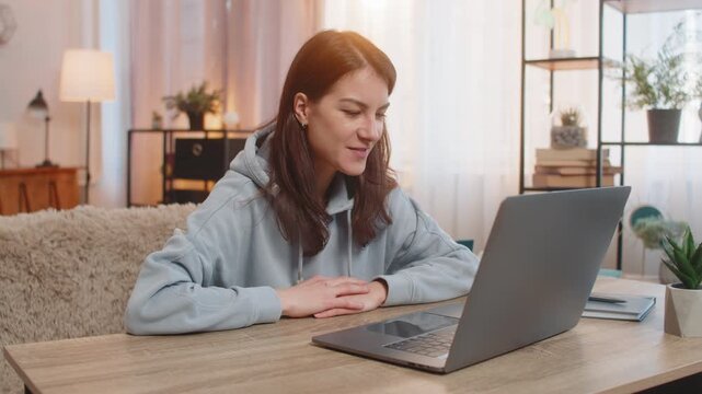 Happy young woman talking to friend by video call using laptop at table smile wave hand share good news looking at screen. Caucasian girl enjoying nice virtual conversation sitting on sofa at home.