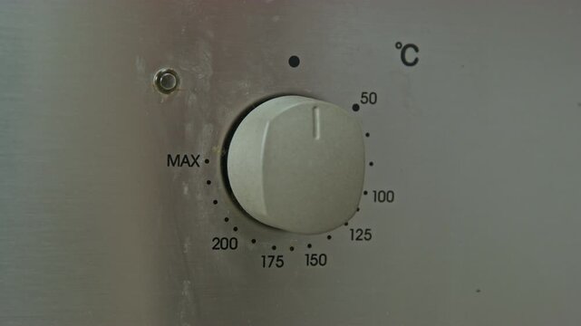 A human hand turns the oven temperature control dial on a stainless steel panel to the MAX mark, demonstrating detailed settings for precise control of the cooking process.