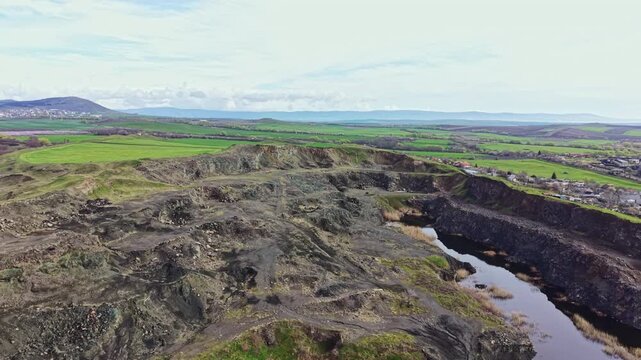 The landscape shows a mining area with rocky terrain and a small lake. Green fields extend in the background, with hills in the distance under a clear sky.