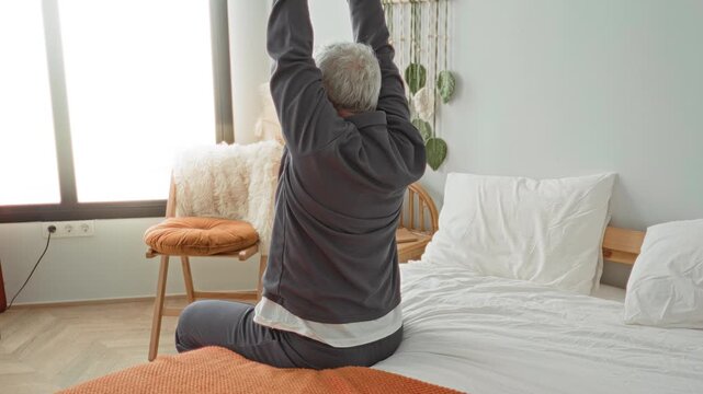 Man on bed stretches arms above head while seated at the mattress edge inside a building; morning calm relaxation.