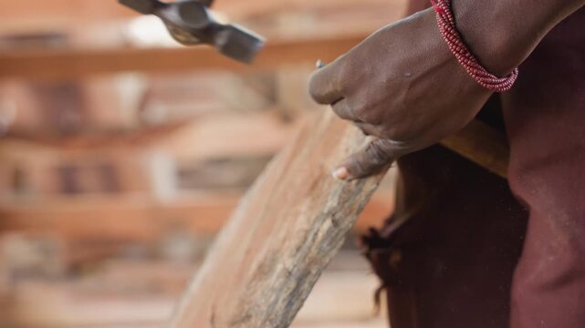 Closeup hands hammering nail into wood, rhythmic strikes creating forceful impact and flying dust, braided bracelet glinting, securing plank joint in sunlit workshop, focused carpentry assembly