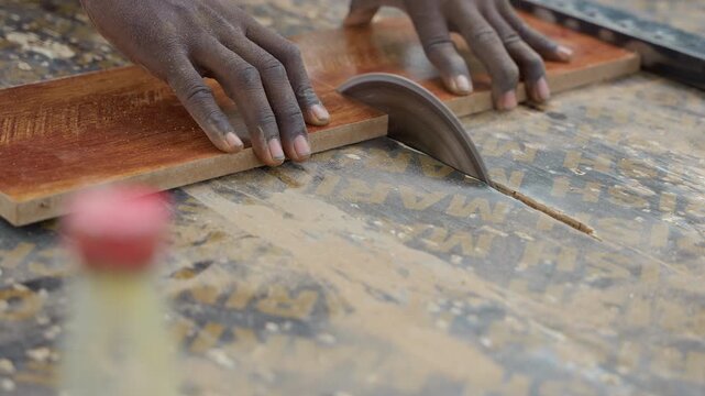 Black hands guiding hardwood across saw, apprentice carpenter trims plank with steady grip, blade slicing through grain, sawdust scattering on workbench, gritty workshop light, focused carpentry craft