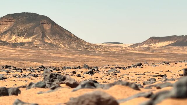 Low angle cinematic panorama of volcanic hill rising above desert plain with basalt rocks in foreground. White Desert National Park, Egypt