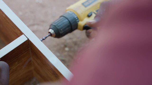 Closeup assembly drilling corner joint, carpenter collects screws in palm after fastening, cordless drill action and steady grip emphasize handwork and technique, wood corner framing and grain