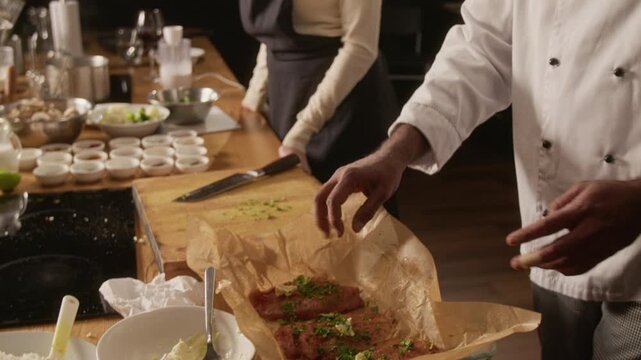 Low angle close up tilt up shot of chef sprinkling herbs over meat in baking dish at counter with rice, spices, and kitchen utensils during group cooking master class in restaurant