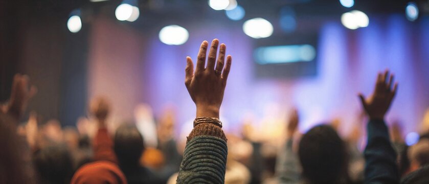 Detail of a hand lifted high in praise during a church service with soft stage lighting and a diverse blurred congregation in the background