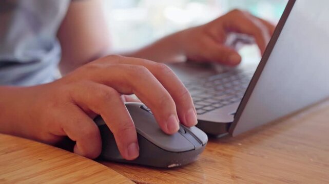 High angle close-up of hands navigating with a computer mouse and typing on a laptop at a wooden table.