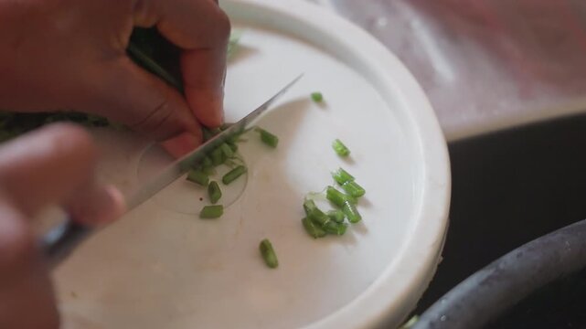 Black hands chopping fresh herbs quickly. Apprentice chef mincing basil and parsley on white cutting board with swift knife motion, small diced garnish forming, focused prep in urban kitchen, precise