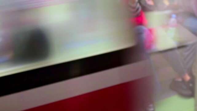 Slow shutter effect of moving commuters move past, blurred and unaware. With passengers visible through glass sits calmly, feet visible.