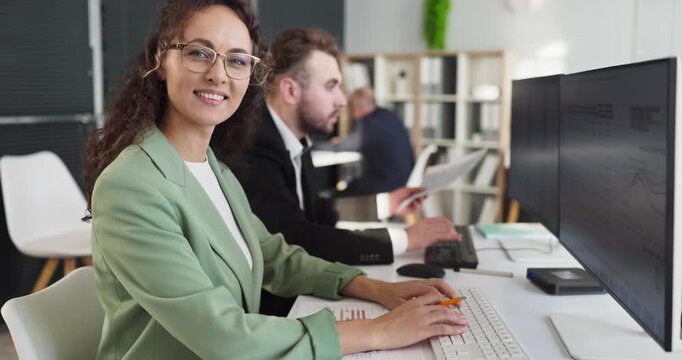 Analyst woman in office at computer. She smiles to camera as colleague reads charts, analyzes data and statistics for finance and report insight on dual screens. Concept of teamwork and analytics.
