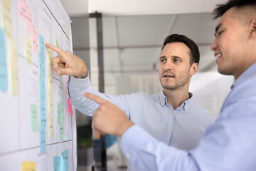 Agile teamwork discussion. Two diverse age multiethnic businessmen colleagues managers executives talk by scrum planning board with colored sticky notes exchange insights review task progress updates © fizkes