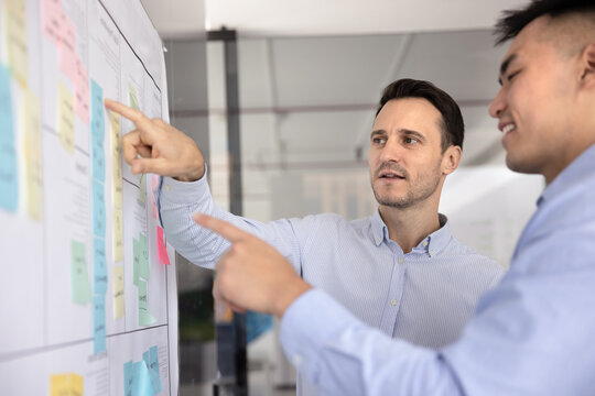 Agile teamwork discussion. Two diverse age multiethnic businessmen colleagues managers executives talk by scrum planning board with colored sticky notes exchange insights review task progress updates